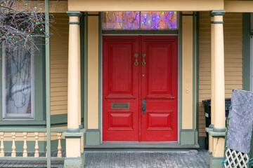 Two tall red solid wood doors with a brass knob, two handles and a letter slot. The historic house is yellow with green trim. There are columns and a veranda with wooden spindles and porch entryway. 