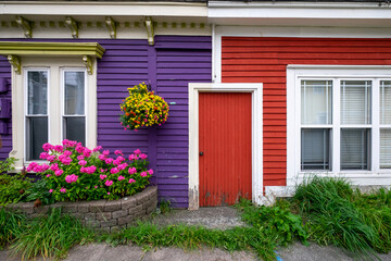 A purple exterior clapboard wooden wall with a hanging flower basket. There's a red door joining the purple wall to a red clapboard wall with a double hung window. There are a flower box and grass too