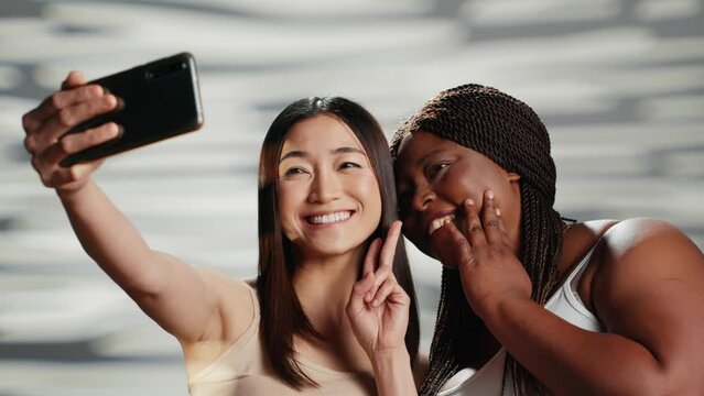 Happy confident women taking photos on smartphone, having fun with pictures on mobile phone in studio. Cheerful silly models being beautiful and posing for body acceptance campaign.