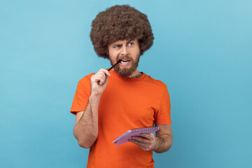Portrait of man with Afro hairstyle in orange T-shirt standing thoughtful biting pencil, pondering...