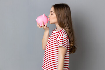 Side view portrait of optimistic little girl wearing striped T-shirt standing holding piggybank, kissing money box. Indoor studio shot isolated on gray background.