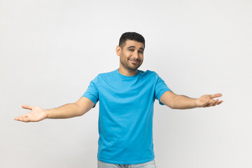 Portrait of unshaven man wearing blue T- shirt standing shows welcome gesture, spreads hands as wants to cuddle best friend, smiles broadly. Indoor studio shot isolated on gray background.