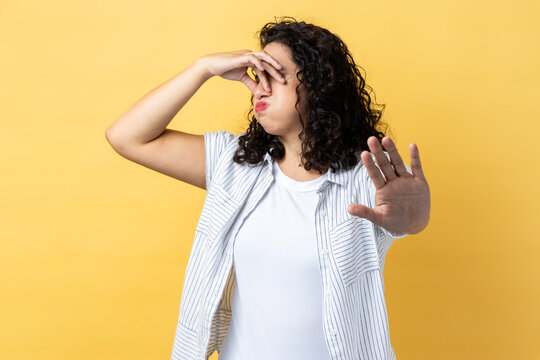 Portrait of woman with dark wavy hair pinching her nose to hold breath and showing stop gesture, disgusted by unpleasant odor, fart. Indoor studio shot isolated on yellow background.