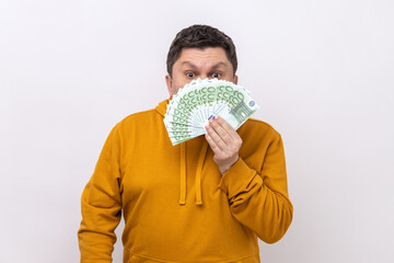 Dark haired man hiding half of face behind fan of hundred euro bills, big winning, betting, wearing...