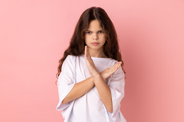 No way, absolutely not. Portrait of little girl wearing white T-shirt showing x sign with crossed hands, meaning stop, this is the end. Indoor studio shot isolated on pink background.