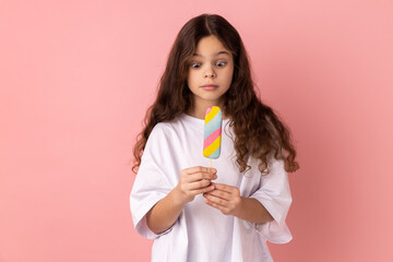 Portrait of shocked surprised little girl wearing white T-shirt holding delicious ice cream, looks at dessert with astonished expression. Indoor studio shot isolated on pink background.