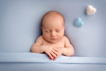 A cute newborn boy in the first days of life sleeps naked on a blue fabric background Light blue and pink felt heart on a blue background. Studio professional macro photography, portrait of a newborn