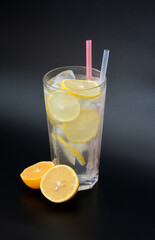 Lemonade, a refreshing drink in a tall glass with ice, straws and slices of ripe lemon on a black background.