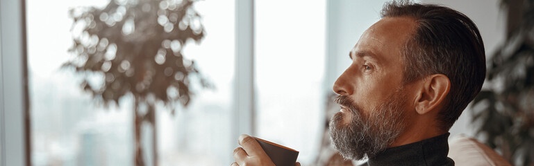 Side view of adult man resting and holding cup of coffee while looking out the window