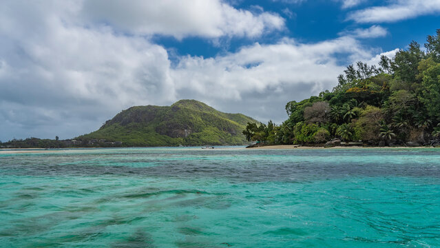 Tropical islands in the turquoise ocean are completely covered with lush vegetation. Boulders on a sandy beach. The boat is visible in the distance. Clouds in the blue sky. Seychelles