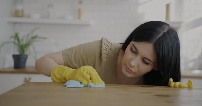 Young woman wearing rubber gloves cleaning kitchen table with wet cloth doing housework alone. Cleanliness and housekeeping activity concept.