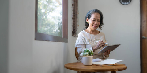 60s aged Asian woman in casual clothes sitting by the window in her living room and using portable digital tablet.