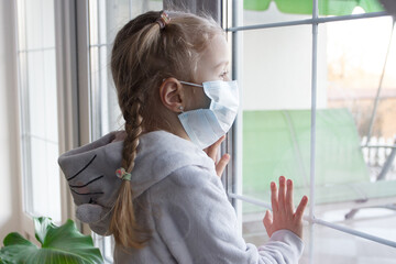 Virus concept, little girl in face mask looking through window at home or clinic. Portrait of sad...