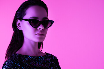 Young beautiful confident brunette female in trendy evening shiny green reflecting dress. Sexy carefree woman isolated on pink neon light in studio. Fashionable model in sunglasses. Fantasy style