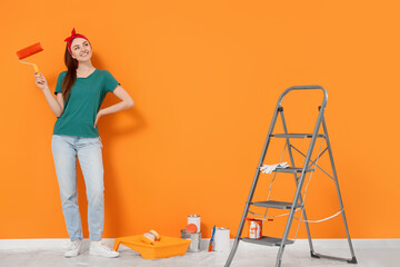 Happy designer with roller and painting equipment near freshly painted orange wall indoors