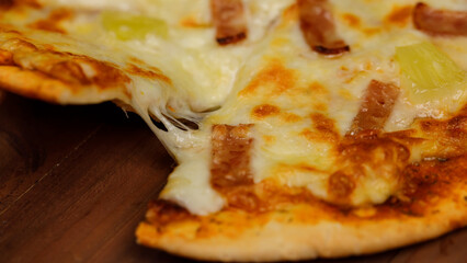 Closeup studio shot using wooden scoop peel picking up hot oven baked tasty delicious Italian homemade ham and pineapple Hawaiian soft bread crust pizza piece from wood plate pan on restaurant table