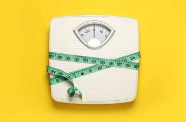 Weigh scales tied with measuring tape on yellow background, top view. Overweight concept