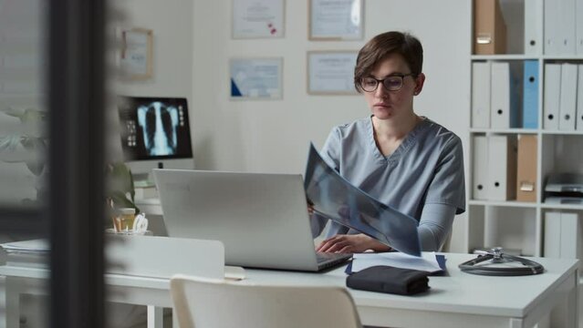 Young serious female clinician in eyeglasses and blue medical scrubs learning x-ray image of patient during online consultation while sitting by workplace in office
