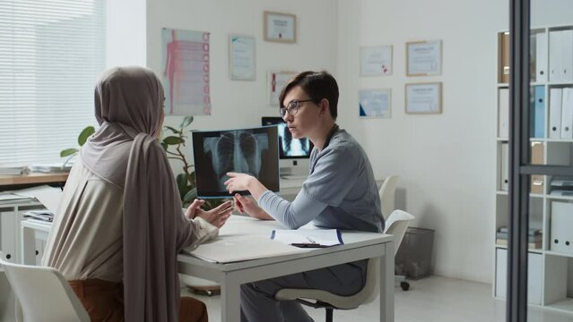 Young confident radiologist with x-ray image listening to Muslim female patient while sitting by workplace in front of her during medical consultation in clinics