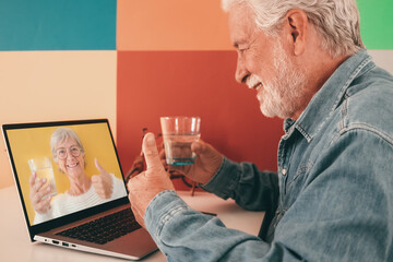 Smiling bearded old senior man chatting in online video using laptop holding a glass of water. Caucasian male having virtual video call with happy wife or friend. Colorful background, copy space