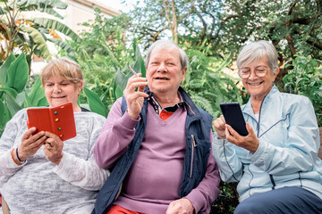 Group of relaxed senior retirees sitting in public park together enjoying free time. Women using phone, man smoking a cigarette