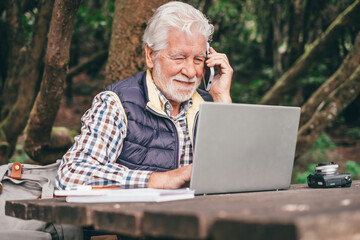 Obraz premium Smiling bearded senior man working on laptop using mobile phone while sitting at a wooden table outdoors in the middle of the park. Concept of smart working in nature, business in fresh air