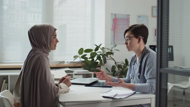 Young Muslim woman in hijab and clinician discussing details of x-ray result while doctor taking it and explaining some points to patient during medical consultation