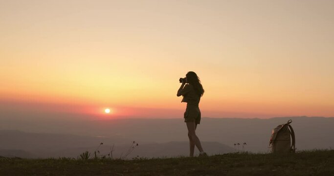 A woman with a professional camera takes a photo of a sunrise in orange and red tones. The woman climbed the very mountain for amazing photos in her portfolio. Silhouette of a young woman photographer