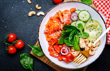 Ketogenic diet salad bowl with salmon, shrimp, avocado, spinach, cucumber, tomato, cashew nuts, sesame. Low-carbohydrate breakfast rich in healthy fats. Black table background, top view