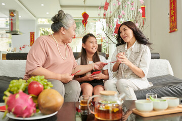 Happy senior woman giving red lucky money envelopes to excited adult daughter and grandaughter