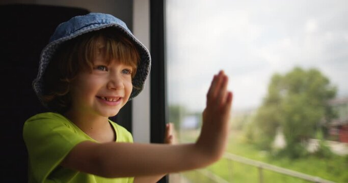 Child smiles and laughs as he looks out train window and sees changing views of surroundings of village. Happy child holds on to glass and looks out the window. Trip to village to visit grandparents 
