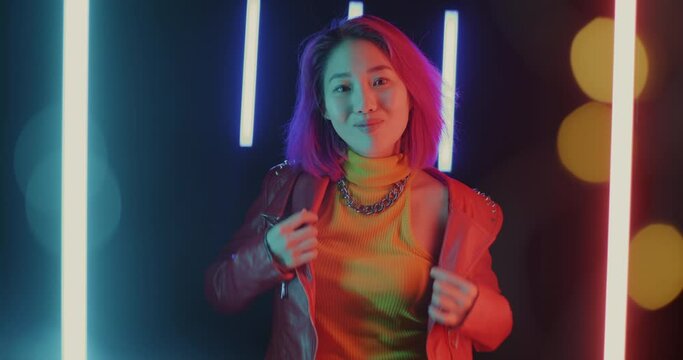 Slow motion portrait of young Asian woman fixing trendy jacket and smiling on futuristic neon background. Fashion and youth concept.