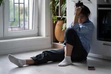 Unhappy depressed Asian girl sitting on floor at home looking at smartphone waiting for call from...