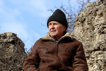 Portrait of elderly man in leather coat and knitted hat in a mountains on blue sky background. Concept of harsh highlander, travel and freedom, healthy old age and adventure on nature