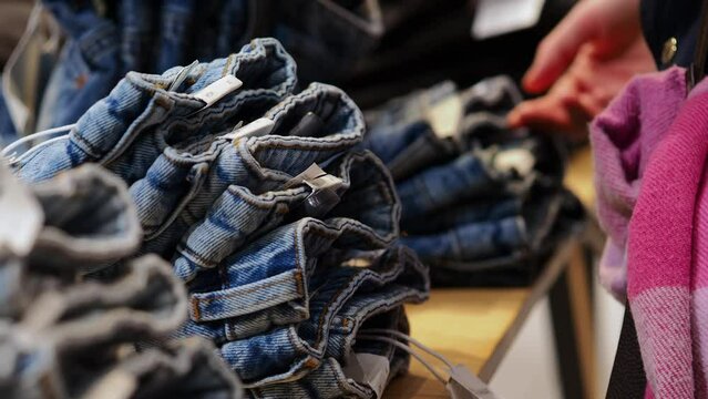 A man in the background is selecting jeans from a display in a retail store. The focus is on neatly stacked denim, showcasing the variety available for shoppers to choose.