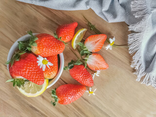 View of whole and halved strawberries inside a white bowl and on top of a wooden table, decorated with lemon slices and flower petals, decorative fabric cloth next to them
