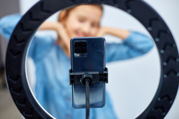 Smiling little girl blogger in casual denim dress use selfie led lamp and smartphone making online video, vlogger live stream isolated on white background, studio. Excited face, lifestyle concept
