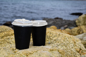 Two paper cups for coffee stand on a stone against the backdrop of the blue sea. Copy space for text. Takeaway cup with lid filled with hot black coffee, milk, ready drink, refreshing.