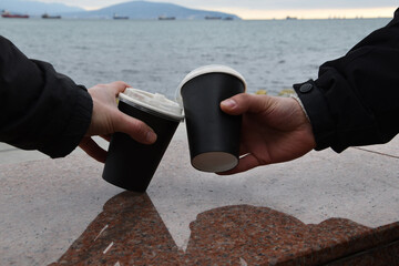 Two paper coffee cups stand on a marble border. Male hands take glasses of coffee against the background of the blue sea. Takeaway cup with lid with ready drink. Recycling of used material, recreation