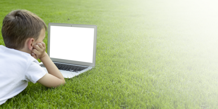 A child sits on the grass and studies with a laptop online, enjoying nature