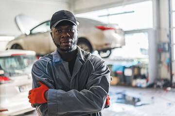 An auto mechanic wearing uniform gloves and a hat stands in a busy auto repair shop. High-quality...