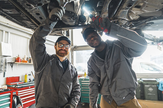 Medium shot of two mechanics smiling at the camera while repairing a car's chassis. Repair shop concept. High quality photo