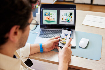 Crop man using smartphone while working on laptop in studio