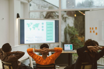 back view of three young colleagues in office sitting with crossed arms behind the neck looking at screen slides studying strategy