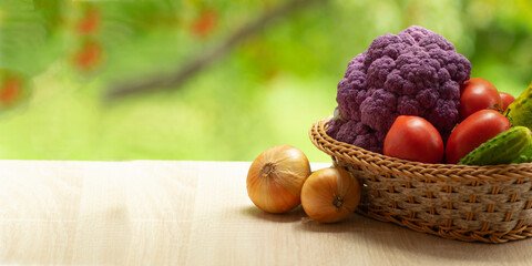 Fresh ripe purple cauliflower, onion, red tomatoes and green cucumber in basket. Healthy food on table on defocus autumn background.