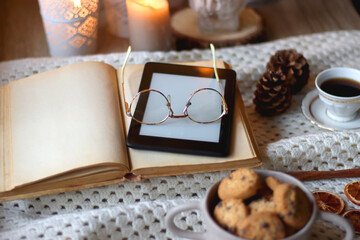 Cup of tea, books, tablet, bowl of cookies, various spices, pine cones and lit candles. Hygge at home. Selective focus.