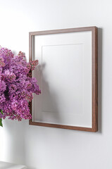 Blank wooden portrait frame mockup with fresh lilac flowers over white wall