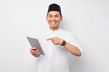 Smiling young Asian Muslim man pointing finger at digital tablet isolated on white background. People religious Islam lifestyle concept. celebration Ramadan and ied Mubarak