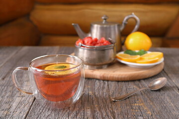 A glass cup of black tea with lemon and mint next to an old iron teapot and other tea accessories stands on a table against a log wall. 