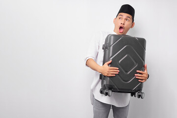 Shocked young Asian Muslim man hugging a suitcase and looking aside isolated on white background. Ramadan and eid Mubarak concept